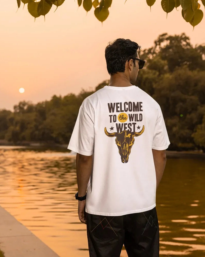 Man wearing a white t-shirt with 'Welcome to the Wild West' design, standing by a lake at sunset.