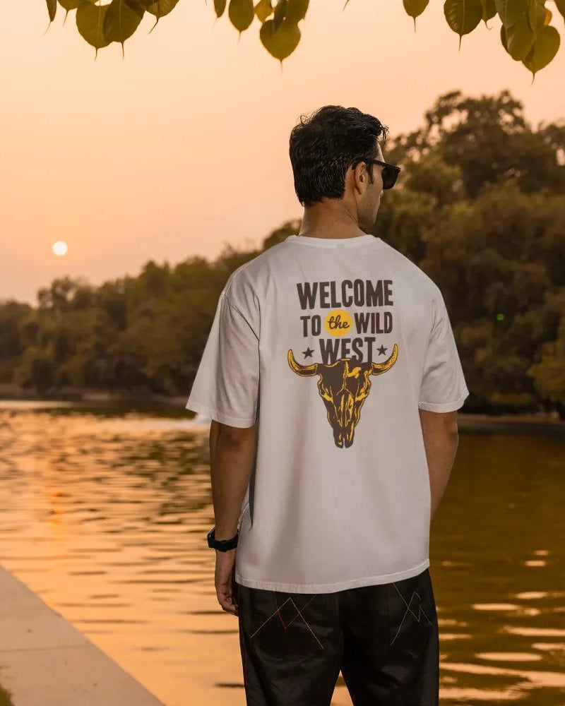 Man wearing a t-shirt with 'Welcome to the Wild West' design by a lake at sunset.